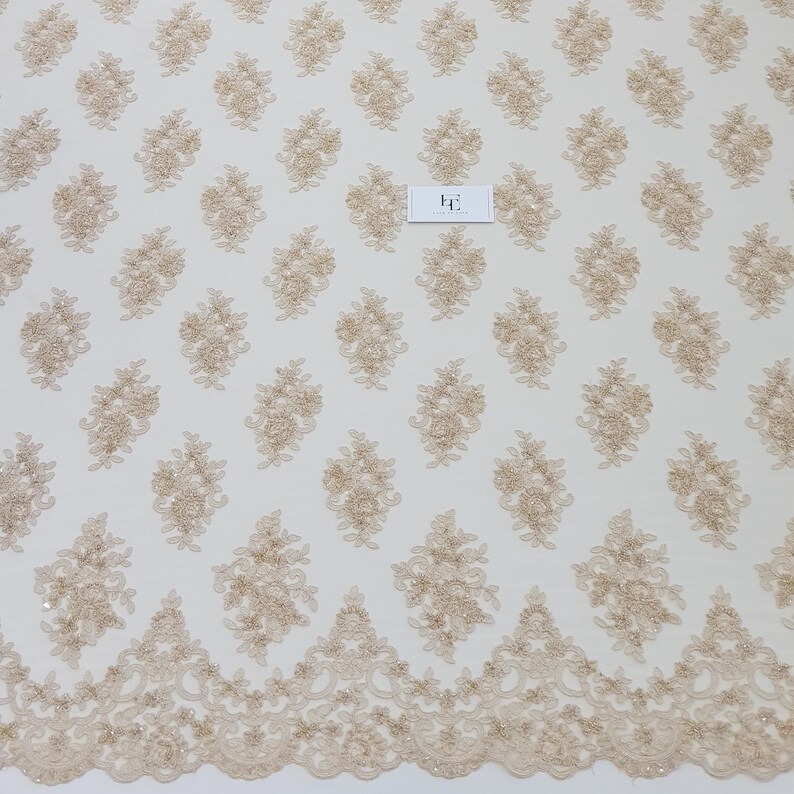 May include: Ivory lace fabric with a repeating floral pattern in a light tan colour. The fabric features a scalloped edge with detailed floral and leaf designs. The lace is suitable for bridal wear, dresses, or other decorative projects.