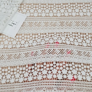 May include: White lace fabric with a delicate floral pattern. The lace has a scalloped edge and is perfect for adding a touch of elegance to any project.