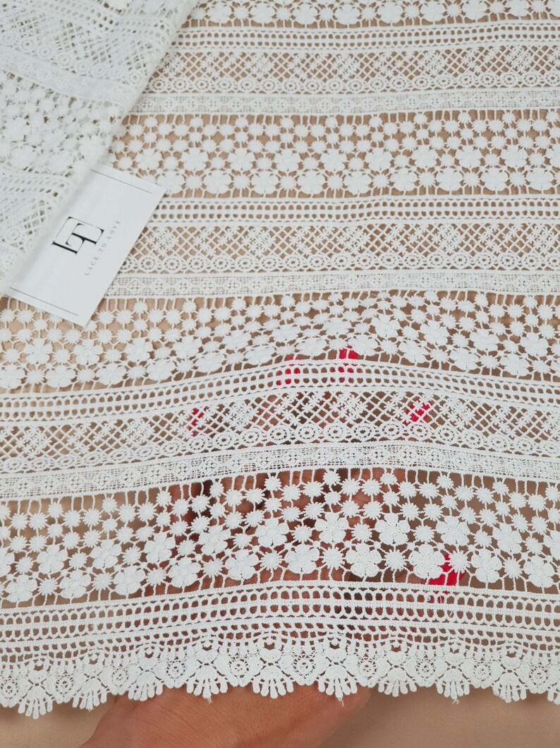May include: White lace fabric with a delicate floral pattern. The lace has a scalloped edge and is perfect for adding a touch of elegance to any project.