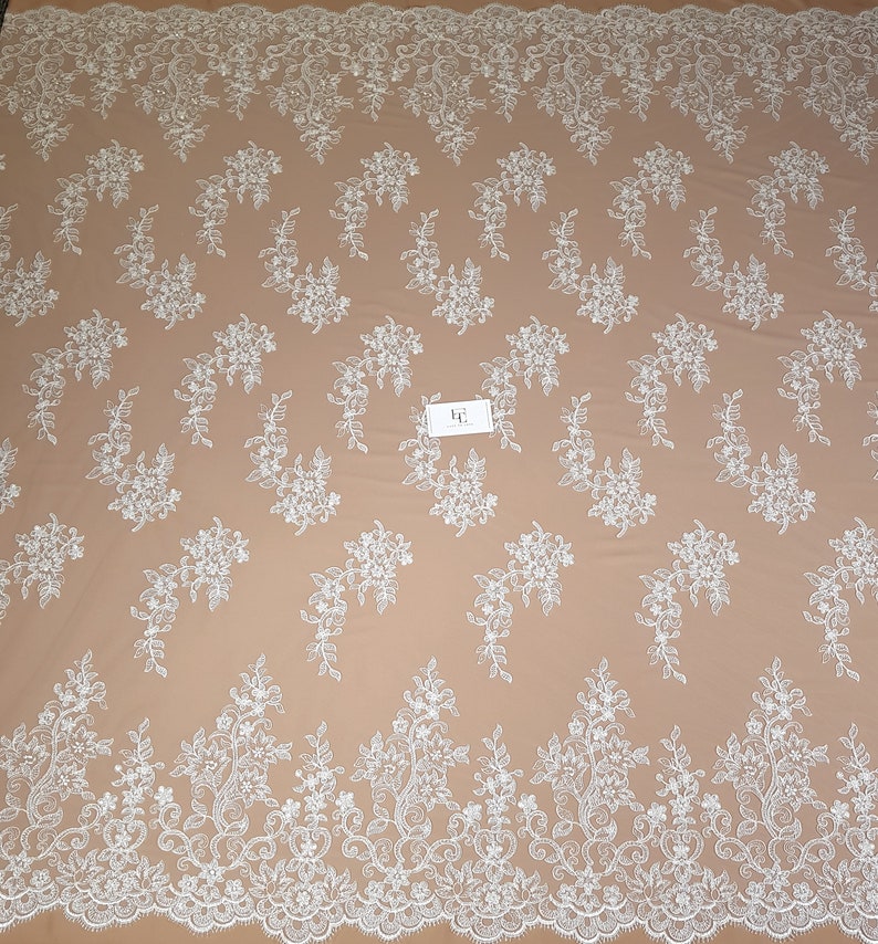 May include: White floral lace fabric with a delicate pattern. The lace has a scalloped edge and is perfect for crafting, sewing, or decorating.