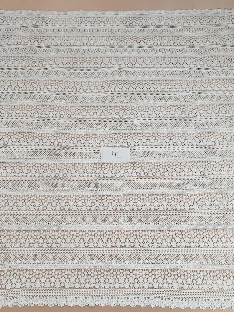 May include: White crochet lace fabric with a repeating pattern of small flowers and geometric shapes. The fabric has a scalloped edge.