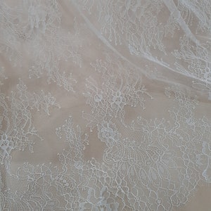 Pode incluir: Close-up de tecido de renda floral branca. A delicada renda apresenta padr&otilde;es intrincados de flores e folhas, criando um design texturizado e elegante. O tecido &eacute; drapeado, revelando os intrincados detalhes da renda.