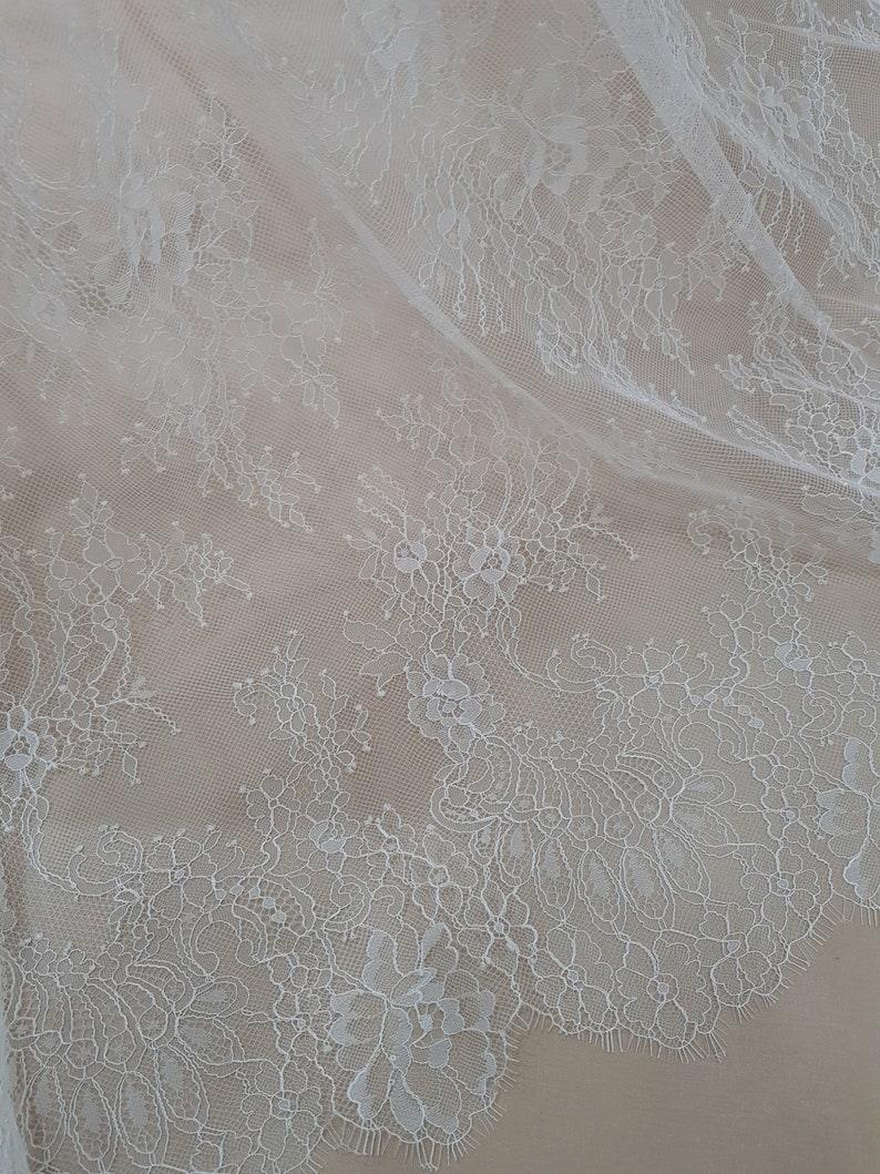 Pode incluir: Close-up de tecido de renda floral branca. A delicada renda apresenta padr&otilde;es intrincados de flores e folhas, criando um design texturizado e elegante. O tecido &eacute; drapeado, revelando os intrincados detalhes da renda.