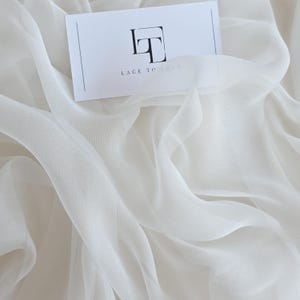 Könnte beinhalten: Cremefarbener, durchscheinender Stoff mit sanften Falten. Eine Visitenkarte mit dem Text "LACE TO LOVE" und einem Logo liegt auf dem Stoff. Der Stoff wirkt leicht und fließend.