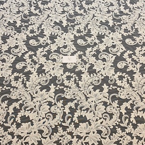 May include: An off-white lace fabric featuring an intricate floral and scrolling vine pattern with scalloped edges. The design includes detailed leaves and blossoms embroidered onto a sheer mesh base, with a small rectangular tag in the center.
