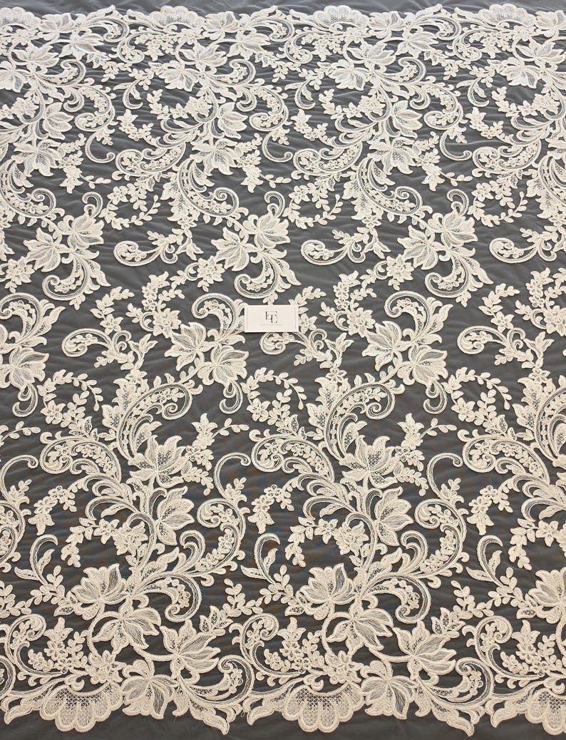 May include: An off-white lace fabric featuring an intricate floral and scrolling vine pattern with scalloped edges. The design includes detailed leaves and blossoms embroidered onto a sheer mesh base, with a small rectangular tag in the center.