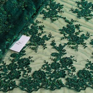 以下が含まれることがあります： エメラルドグリーンのレース生地。花柄の刺繍とビーズのアクセントが施されています。繊細なデザインは、花と葉の複雑なパターンを特徴とし、小さく丸いビーズが質感と奥行きを加えています。生地は半透明で、柔らかく流れるようなドレープです。