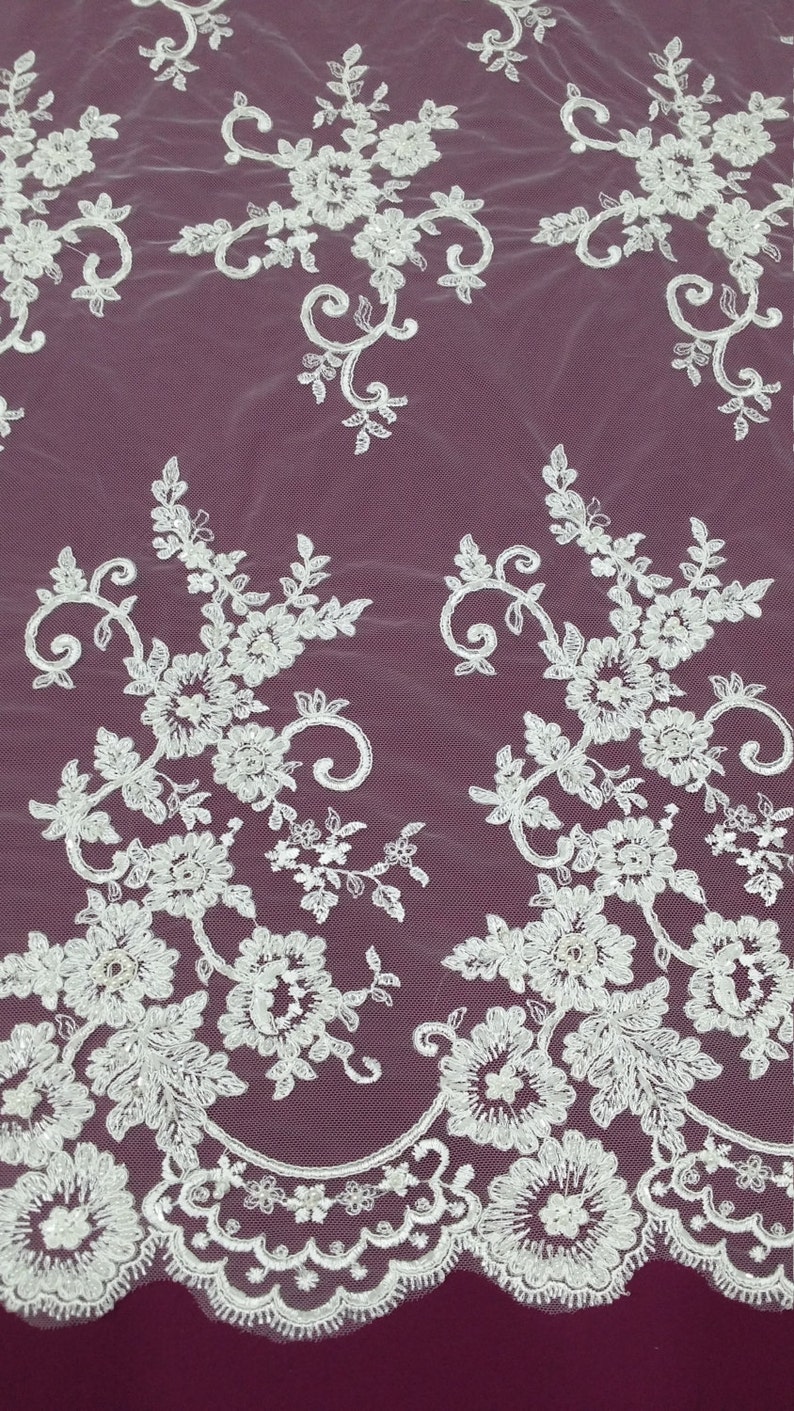 May include: White floral lace fabric with detailed embroidered flowers and leaves. The lace features a delicate, open weave design with a scalloped edge. The fabric is set against a deep burgundy background, creating a striking contrast.