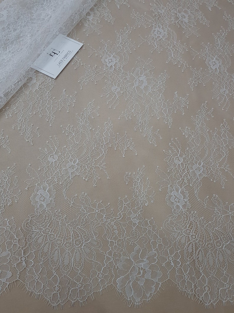 Pode incluir: Tecido de renda floral branca com borda ondulada. A renda apresenta um delicado padr&atilde;o floral e est&aacute; parcialmente enrolada. Uma etiqueta com o texto "Lace Fabrics" &eacute; vis&iacute;vel.