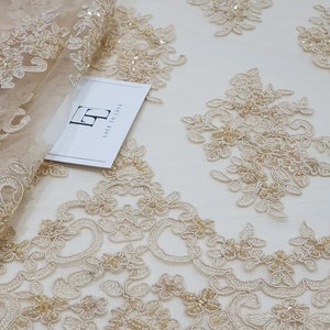 May include: Close-up of beige lace fabric with detailed floral embroidery and bead embellishments. The lace has a delicate pattern of flowers and swirling designs, suitable for bridal wear or special occasion garments. The fabric is soft and sheer, with a subtle sheen.