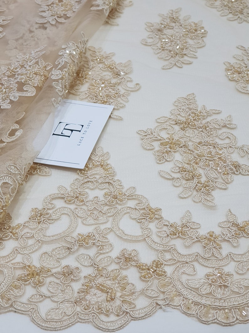 May include: Close-up of beige lace fabric with detailed floral embroidery and bead embellishments. The lace has a delicate pattern of flowers and swirling designs, suitable for bridal wear or special occasion garments. The fabric is soft and sheer, with a subtle sheen.