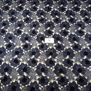 May include: A navy blue lace fabric featuring a repeating pattern of embroidered flowers with textured centres and detailed leaves. A small white rectangular tag with the black text "HE" is positioned in the centre of the floral design.