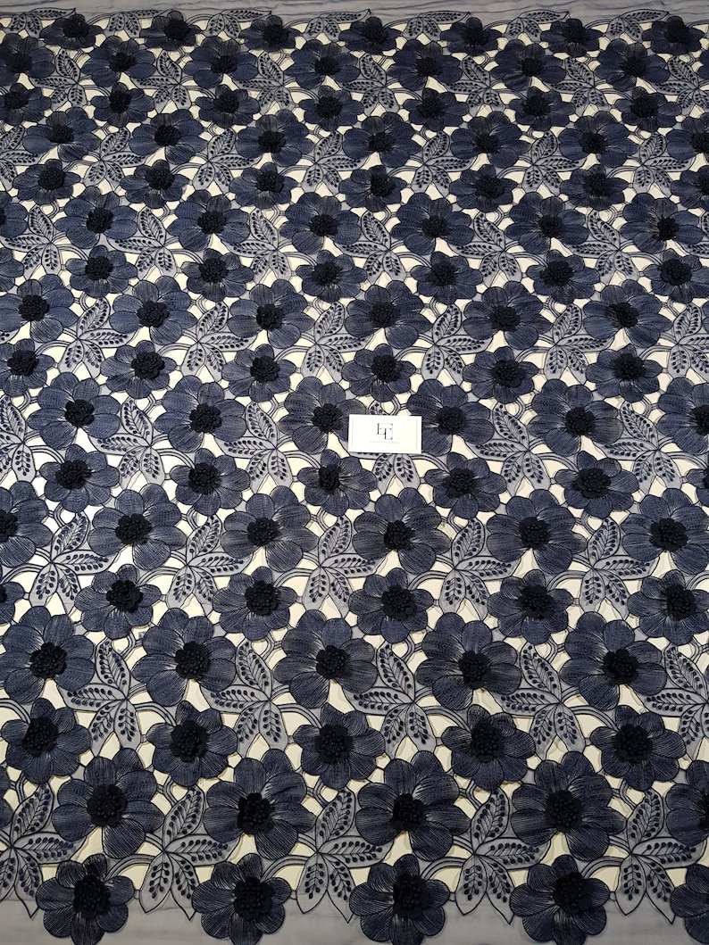 May include: A navy blue lace fabric featuring a repeating pattern of embroidered flowers with textured centres and detailed leaves. A small white rectangular tag with the black text "HE" is positioned in the centre of the floral design.