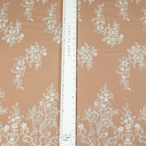 May include: White floral lace fabric with a scalloped edge. The fabric is on a peach-colored background. A ruler is visible on the right side of the image. The ruler is marked in inches and centimeters. The ruler reads "Lace to Love" and has a measurement of 30 inches.