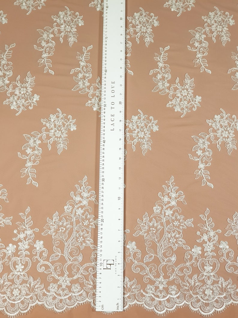 May include: White floral lace fabric with a scalloped edge. The fabric is on a peach-colored background. A ruler is visible on the right side of the image. The ruler is marked in inches and centimeters. The ruler reads "Lace to Love" and has a measurement of 30 inches.