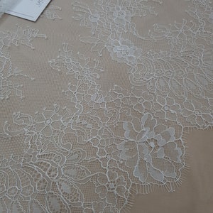Pode incluir: Close-up de tecido de renda floral branca com borda ondulada. A renda apresenta padr&otilde;es detalhados de flores e folhas. Uma etiqueta com o texto "LACE FABRICS" &eacute; vis&iacute;vel no canto superior esquerdo. O tecido &eacute; exibido em um fundo bege.