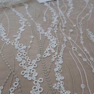 May include: A sheer beige fabric featuring white embroidered patterns of vertical, flowing vine-like lines. The embroidery consists of small circular shapes and delicate, thin metallic silver threads that create a textured, organic design across the surface.