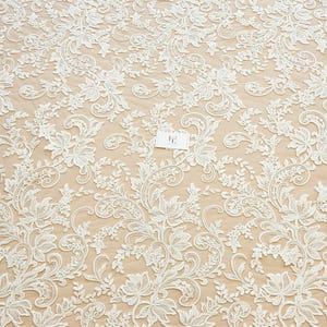 May include: An off-white lace fabric featuring an intricate floral and vine pattern with embroidered details. The lace is layered over a light beige background and includes a small white rectangular tag with the letters FE printed in black.