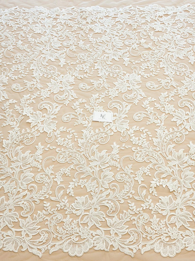 May include: An off-white lace fabric featuring an intricate floral and vine pattern with embroidered details. The lace is layered over a light beige background and includes a small white rectangular tag with the letters FE printed in black.