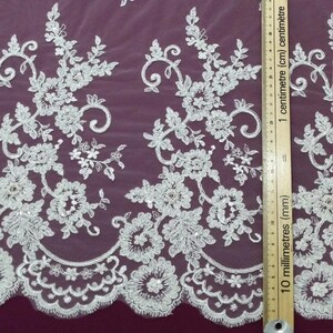May include: White floral lace fabric with intricate embroidered details. The design features flowers, leaves, and swirling patterns. The lace is measured with a ruler in centimeters.