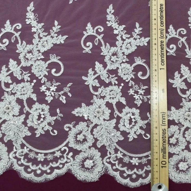 May include: White floral lace fabric with intricate embroidered details. The design features flowers, leaves, and swirling patterns. The lace is measured with a ruler in centimeters.