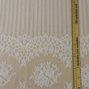 May include: White lace fabric with a floral pattern and a striped background. The fabric is measured with a ruler in inches and centimeters.