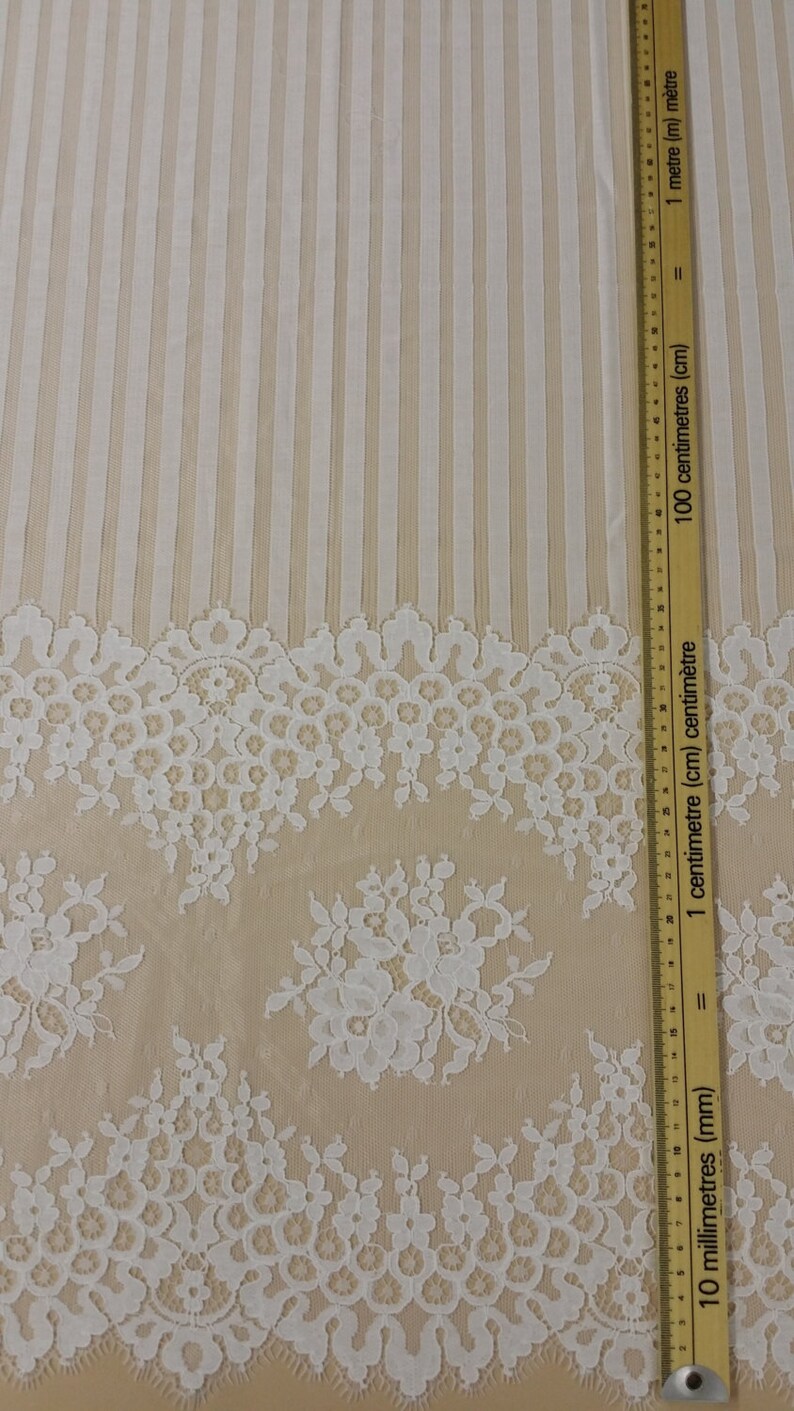May include: White lace fabric with a floral pattern and a striped background. The fabric is measured with a ruler in inches and centimeters.