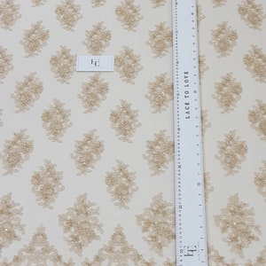 May include: Beige lace fabric with a repeating floral pattern and a scalloped edge. The fabric features embroidered floral designs with sequins. A ruler with both inches and centimetres is visible, along with a small tag that reads "Lace to Love".