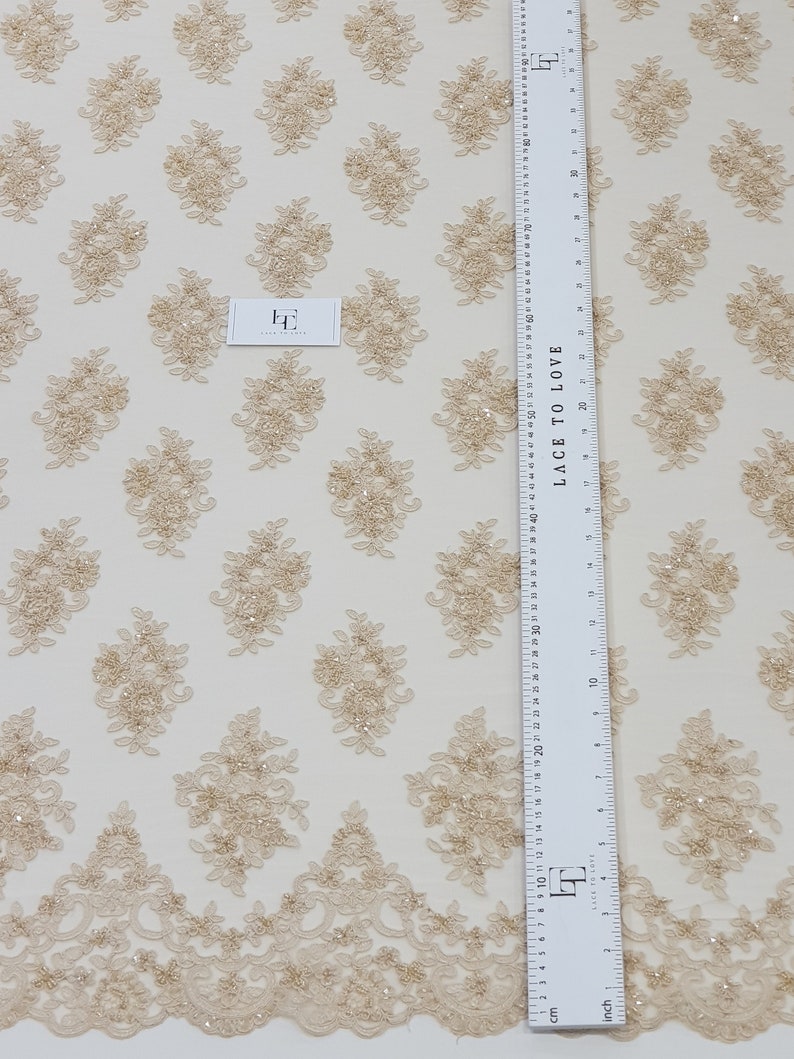 May include: Beige lace fabric with a repeating floral pattern and a scalloped edge. The fabric features embroidered floral designs with sequins. A ruler with both inches and centimetres is visible, along with a small tag that reads "Lace to Love".