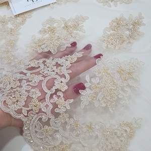 May include: Ivory lace fabric with floral embroidery and beaded accents. The delicate design features intricate patterns of flowers and leaves, with small beads adding texture and dimension. The fabric is ideal for bridal wear and special occasion garments.