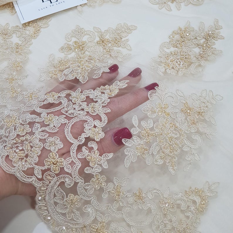 May include: Ivory lace fabric with floral embroidery and beaded accents. The delicate design features intricate patterns of flowers and leaves, with small beads adding texture and dimension. The fabric is ideal for bridal wear and special occasion garments.