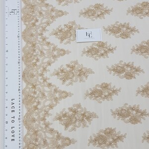 May include: Beige lace fabric with floral embroidery and beaded accents. The fabric features a repeating pattern of embroidered floral clusters and a decorative border. A ruler with centimetre measurements is visible. The tag reads "LACE TO LOVE".
