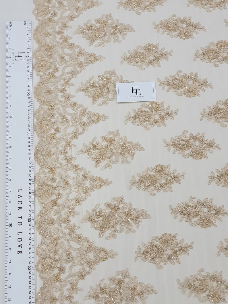 May include: Beige lace fabric with floral embroidery and beaded accents. The fabric features a repeating pattern of embroidered floral clusters and a decorative border. A ruler with centimetre measurements is visible. The tag reads "LACE TO LOVE".