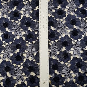 May include: A navy blue floral lace fabric featuring an intricate embroidered pattern of flowers and leaves. A white ruler is placed vertically over the fabric, showing measurements in inches and centimetres, with the text LACE TO LOVE printed on it.