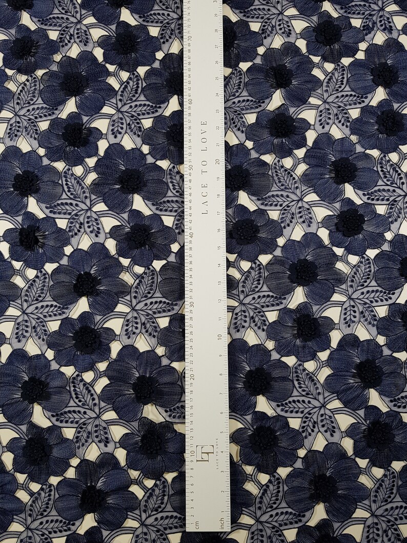 May include: A navy blue floral lace fabric featuring an intricate embroidered pattern of flowers and leaves. A white ruler is placed vertically over the fabric, showing measurements in inches and centimetres, with the text LACE TO LOVE printed on it.