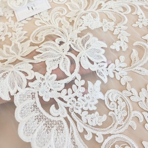 May include: A sheer off-white tulle fabric featuring intricate white floral embroidery with scrolling vine patterns. The delicate lace design includes detailed petals and leaves, creating a continuous botanical motif across the mesh material.