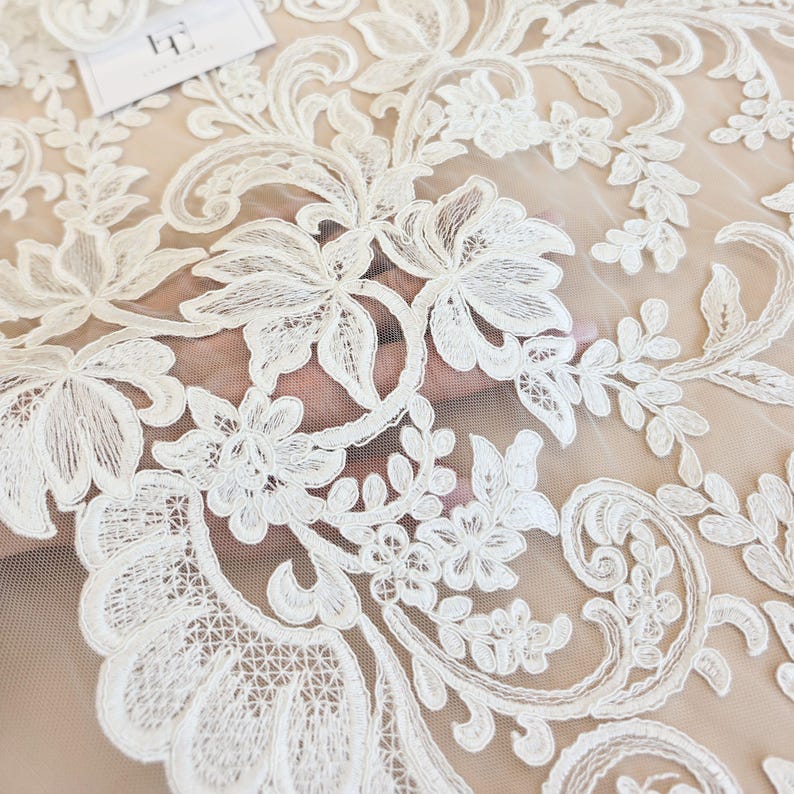May include: A sheer off-white tulle fabric featuring intricate white floral embroidery with scrolling vine patterns. The delicate lace design includes detailed petals and leaves, creating a continuous botanical motif across the mesh material.