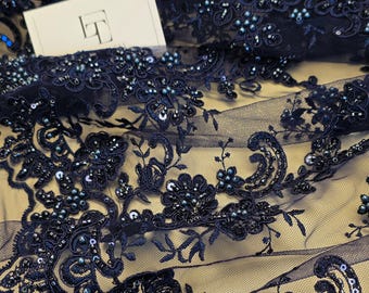May include: Navy blue lace fabric featuring intricate floral embroidery, embellished with matching dark beads and sequins. A small white rectangular tag with the black letters LT is visible in the upper left corner of the sheer mesh material.