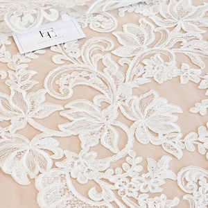 May include: White floral lace fabric featuring intricate embroidered patterns on a sheer nude mesh base. A small white rectangular tag with the black text "LT LACE TO LOVE" is placed on the fabric. The design includes swirling vines and petals.