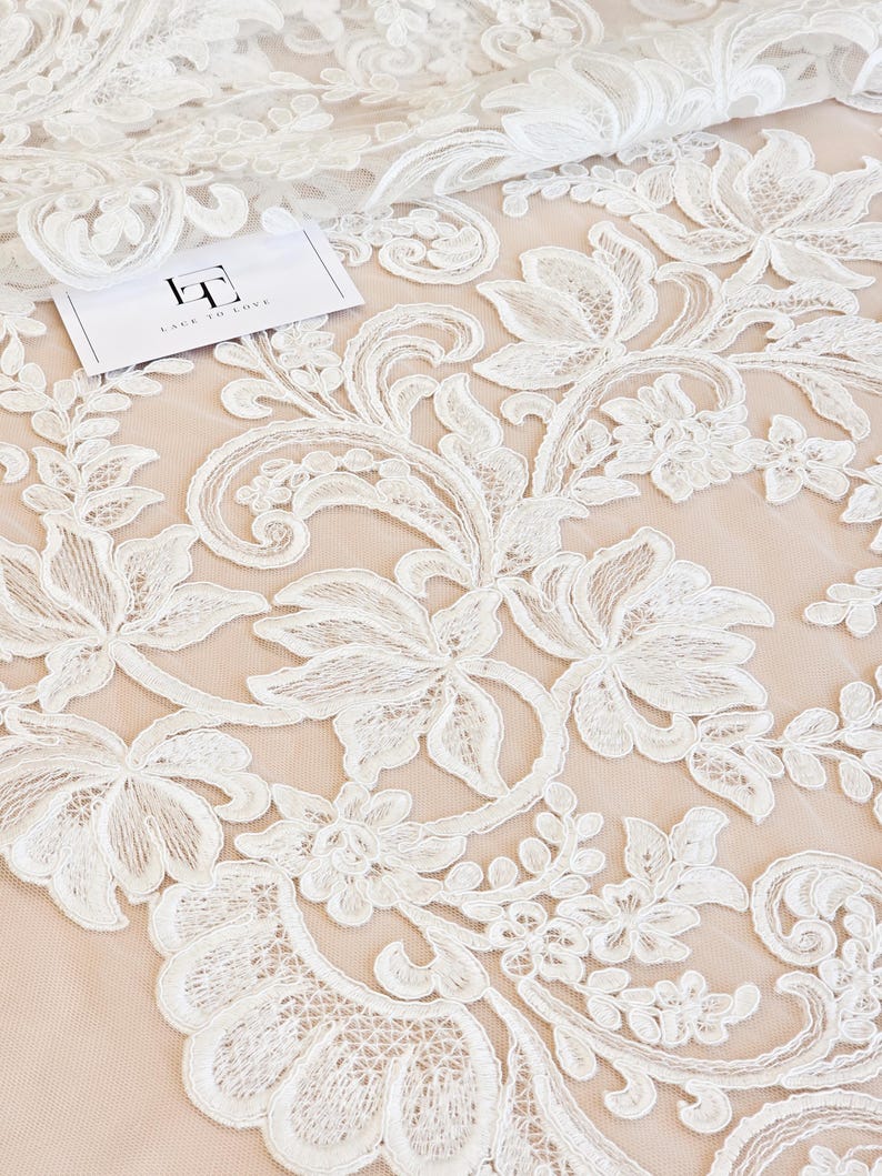 May include: White floral lace fabric featuring intricate embroidered patterns on a sheer nude mesh base. A small white rectangular tag with the black text "LT LACE TO LOVE" is placed on the fabric. The design includes swirling vines and petals.