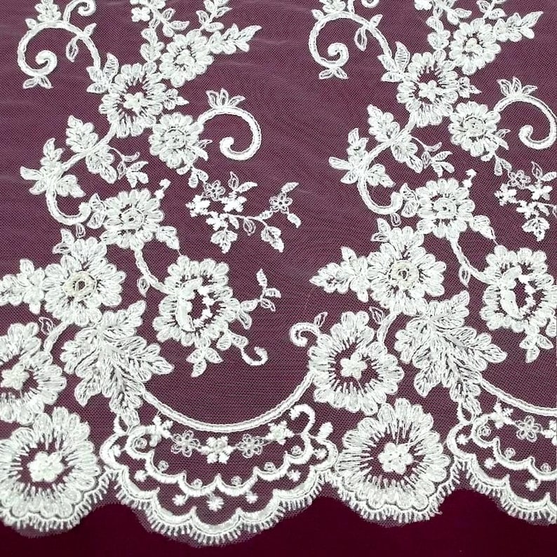 May include: Close-up of white floral lace fabric with intricate embroidered flower and leaf patterns. The lace is set against a deep burgundy background, showcasing the delicate details and textures. The design features a scalloped edge.