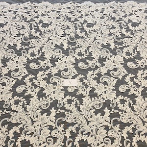 May include: An off-white lace fabric featuring an intricate floral and vine pattern with swirling scrollwork. The design is symmetrical and repetitive, set against a dark background. A small rectangular tag with the letters EF is visible.