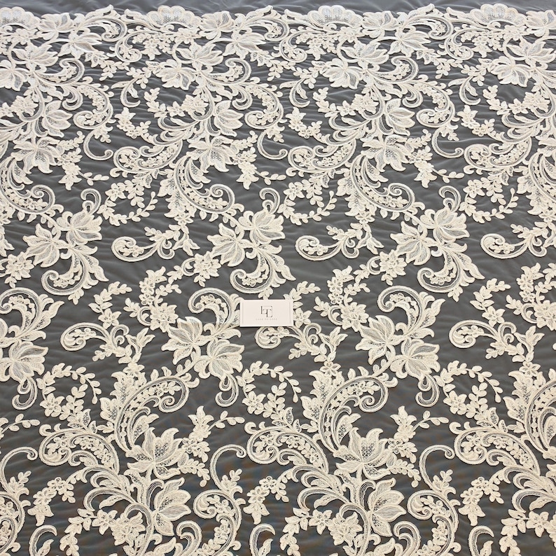 May include: An off-white lace fabric featuring an intricate floral and vine pattern with swirling scrollwork. The design is symmetrical and repetitive, set against a dark background. A small rectangular tag with the letters EF is visible.
