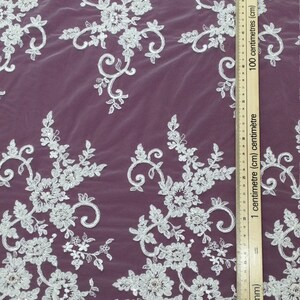 May include: White floral lace fabric with intricate embroidered details. The lace features a repeating pattern of flowers, leaves, and swirling designs. A ruler with centimeter and millimeter markings is placed on the fabric. The fabric is on a dark purple background.