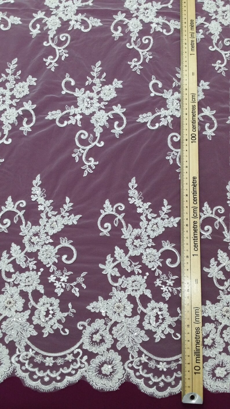 May include: White floral lace fabric with intricate embroidered details. The lace features a repeating pattern of flowers, leaves, and swirling designs. A ruler with centimeter and millimeter markings is placed on the fabric. The fabric is on a dark purple background.