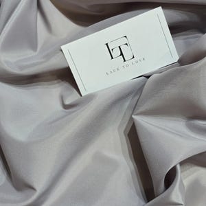 Può includere: Primo piano di un tessuto grigio chiaro con un cartoncino bianco sopra. Il cartoncino riporta il testo "Lace to Love" e un logo con le lettere "LT" in un quadrato.