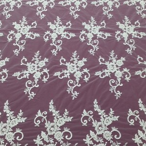 May include: White floral lace fabric with a repeating pattern of flowers and leaves. The lace has a scalloped edge and is set against a burgundy background. This delicate fabric is suitable for bridal wear and other special occasion garments.
