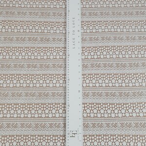 May include: White lace fabric with a repeating pattern of small flowers and geometric designs. The fabric is measured in inches and centimeters.