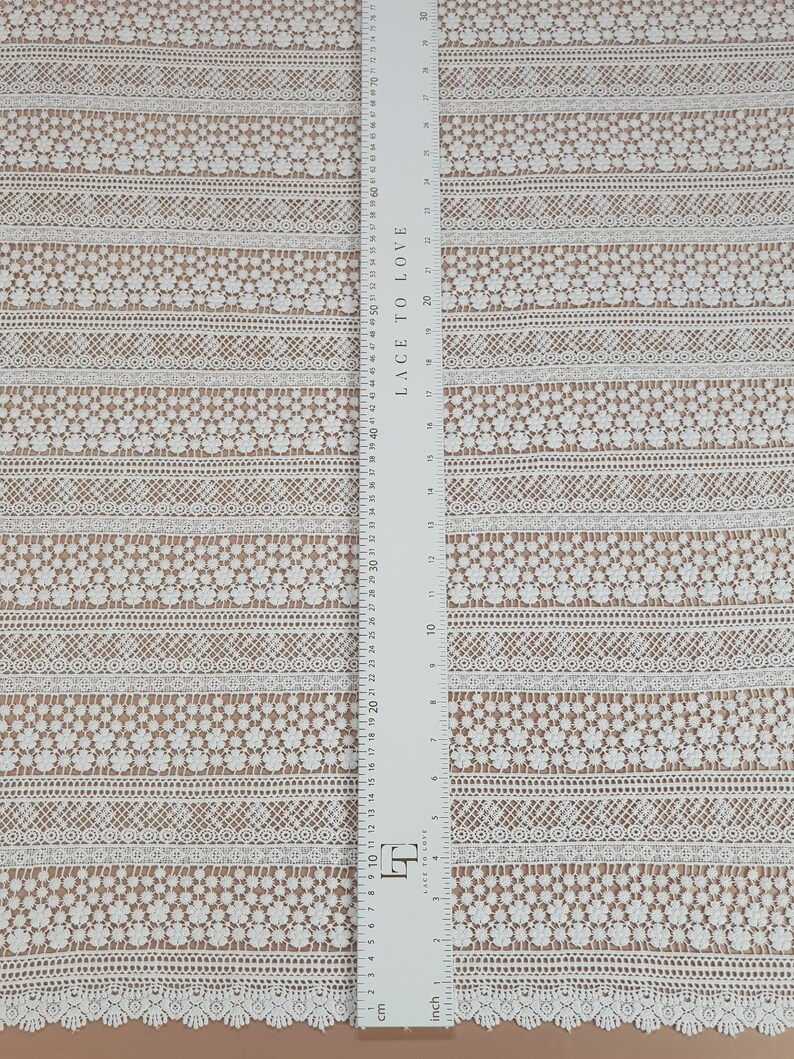 May include: White lace fabric with a repeating pattern of small flowers and geometric designs. The fabric is measured in inches and centimeters.