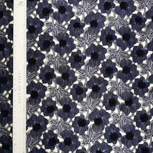 May include: A navy blue floral lace fabric featuring a repeating pattern of embroidered flowers and leaves. A white ruler is placed along the left side, showing a length of 92 centimetres. The lace has a delicate, openwork design with textured centres.